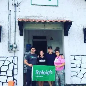 A group of four Raleigh alumni smiling and holding a green Raleigh flag in front of the fieldbase entrance.