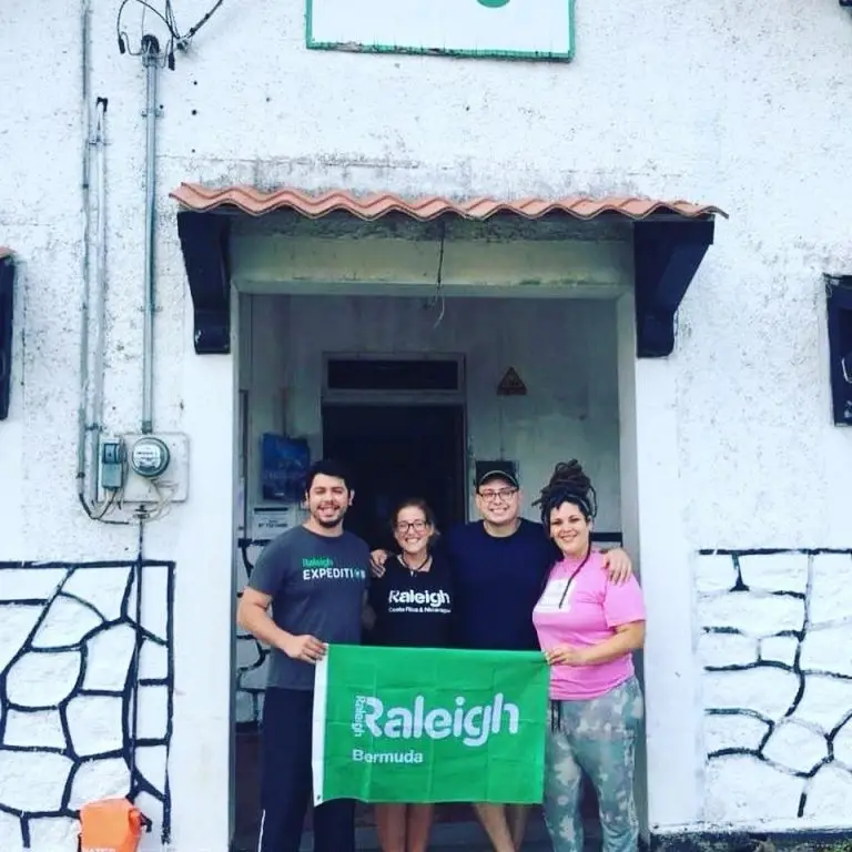 A group of four Raleigh alumni smiling and holding a green Raleigh flag in front of the fieldbase entrance.